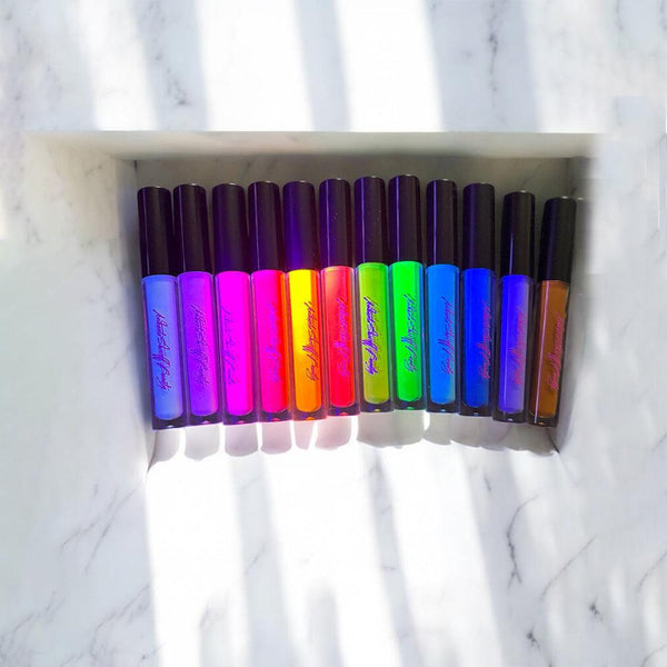 A row of eleven vibrant, UV-reactive vegan paint liners in assorted bold colors arranged on a white marble surface.