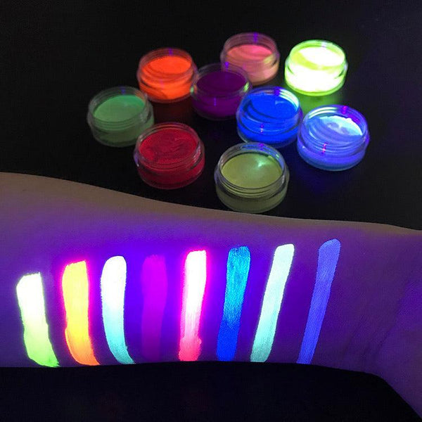 Eight small jars of vibrant neon UV face and body paint with matching colorful swatches glowing on an arm under blacklight.